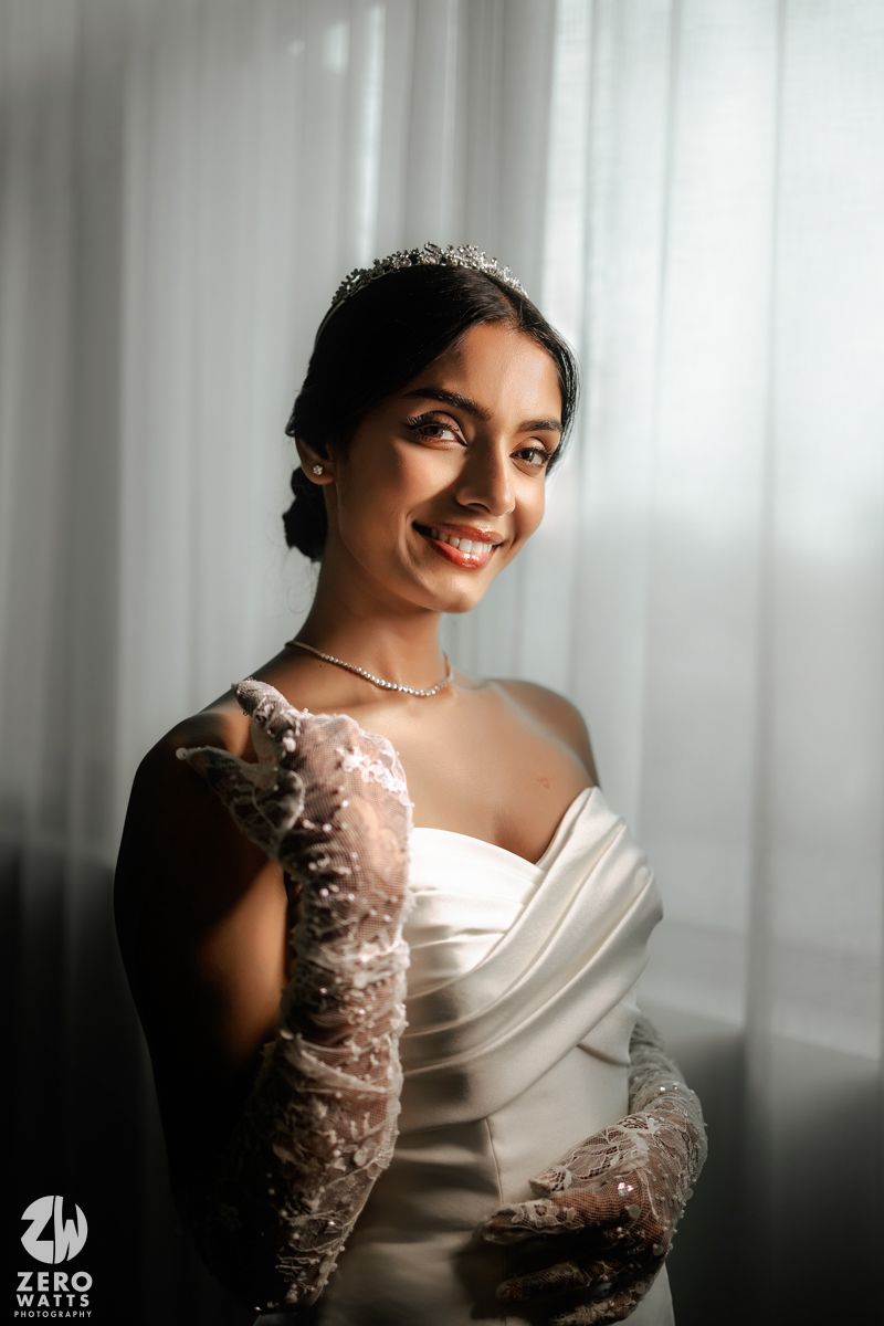 Radiant bride in white dress with lace gloves and tiara smiling softly.