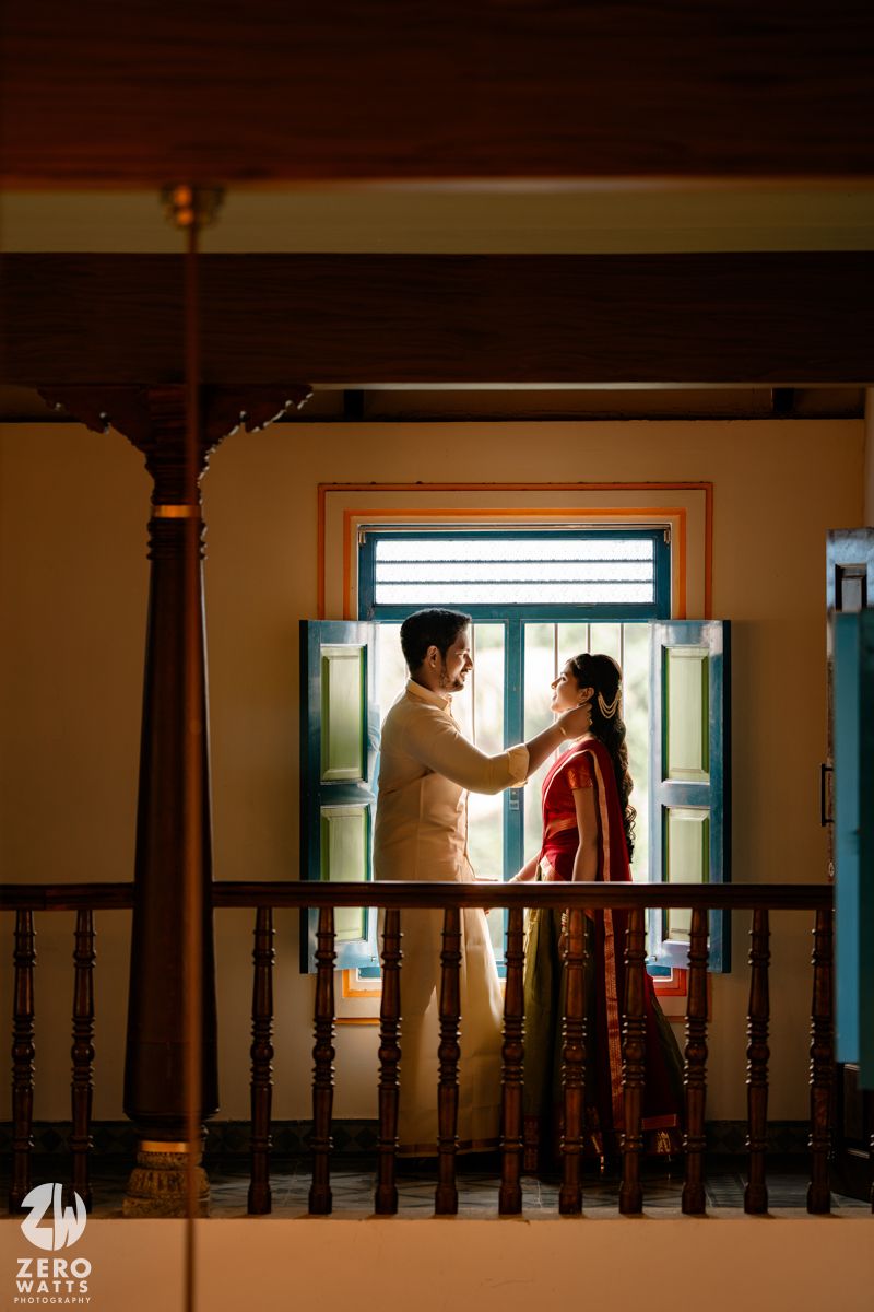 Couple sharing a romantic moment on a balcony, bride in red saree and groom in cream kurta, framed by wooden pillars.