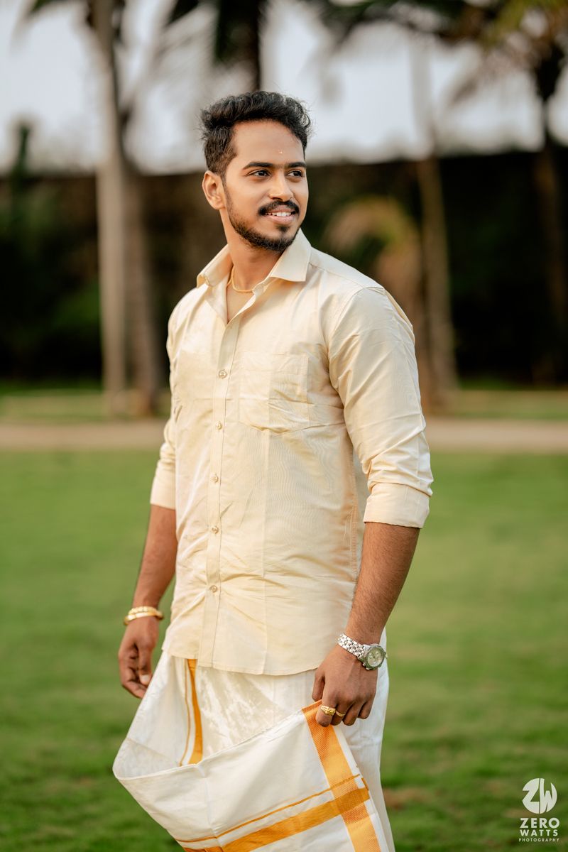 South Indian groom in traditional cream silk shirt and white dhoti with gold border stands outdoors.