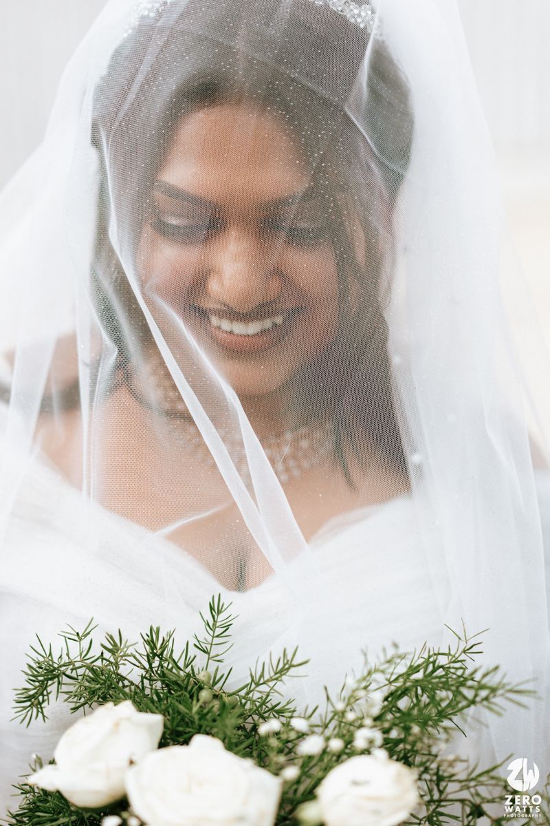 Smiling Indian bride under white veil holding bouquet of white roses and greenery on her wedding day.
