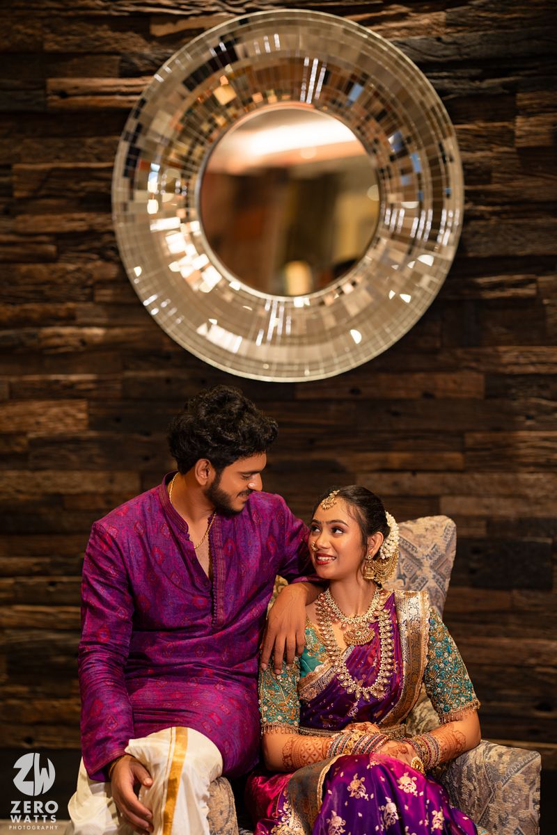 South Indian wedding couple in matching purple outfits sitting together beneath decorative mirror.