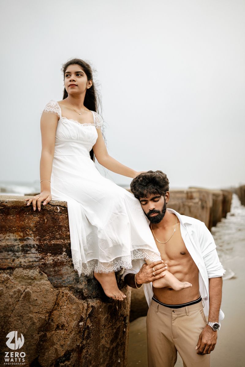 Couple in white outfits posing on rocky sea wall, woman in lace dress seated above man in open shirt at beach.
