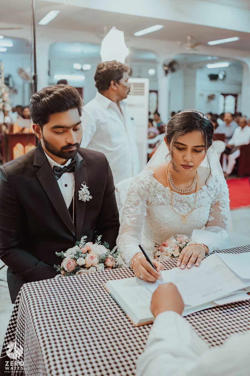 Wedding ceremony signing with couple at checkered table and priest in white robe standing behind.