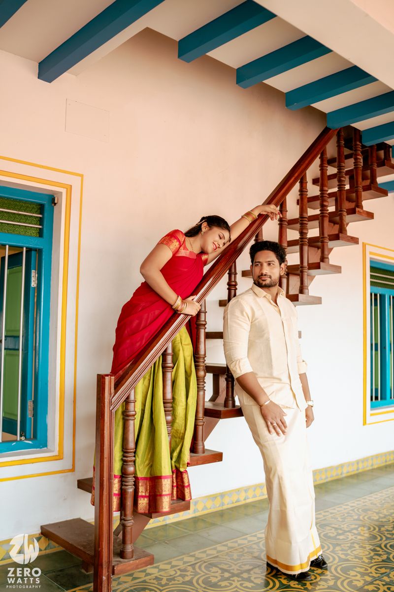 Couple in traditional South Indian attire posing on wooden staircase, woman in red-green saree, man in cream dhoti.