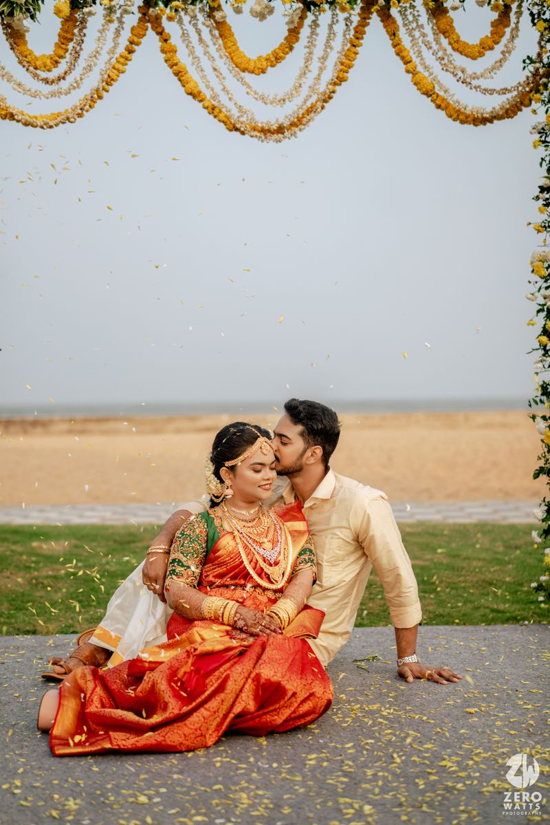 South Indian groom kissing bride in red Kanjivaram saree with gold jewelry under marigold garlands by the beach.