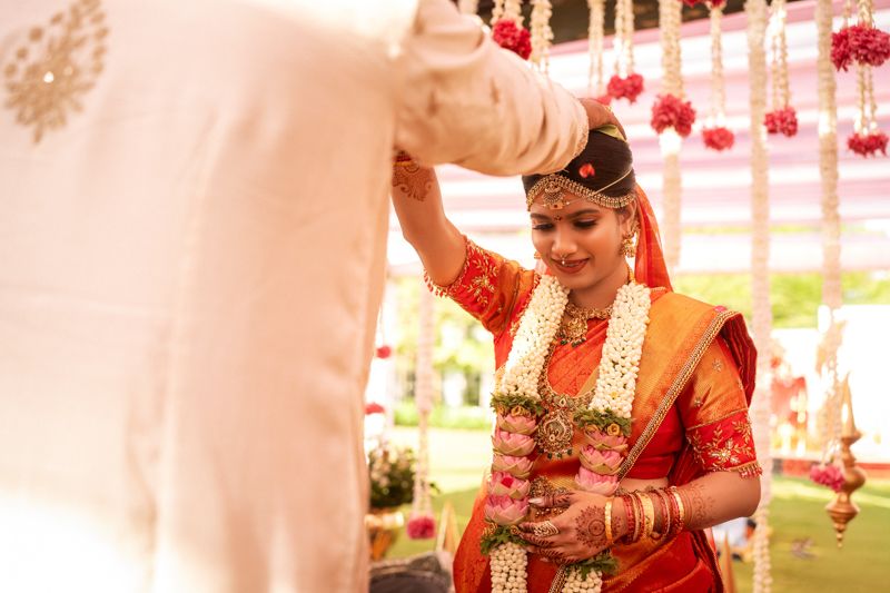 Bride in red saree receiving garland during traditional South Indian wedding ceremony.