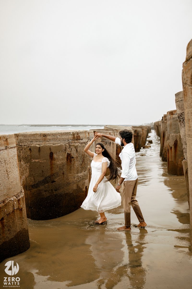 Couple dancing barefoot on a wet beach between stone pillars, woman in white dress, man in white shirt and brown pants.