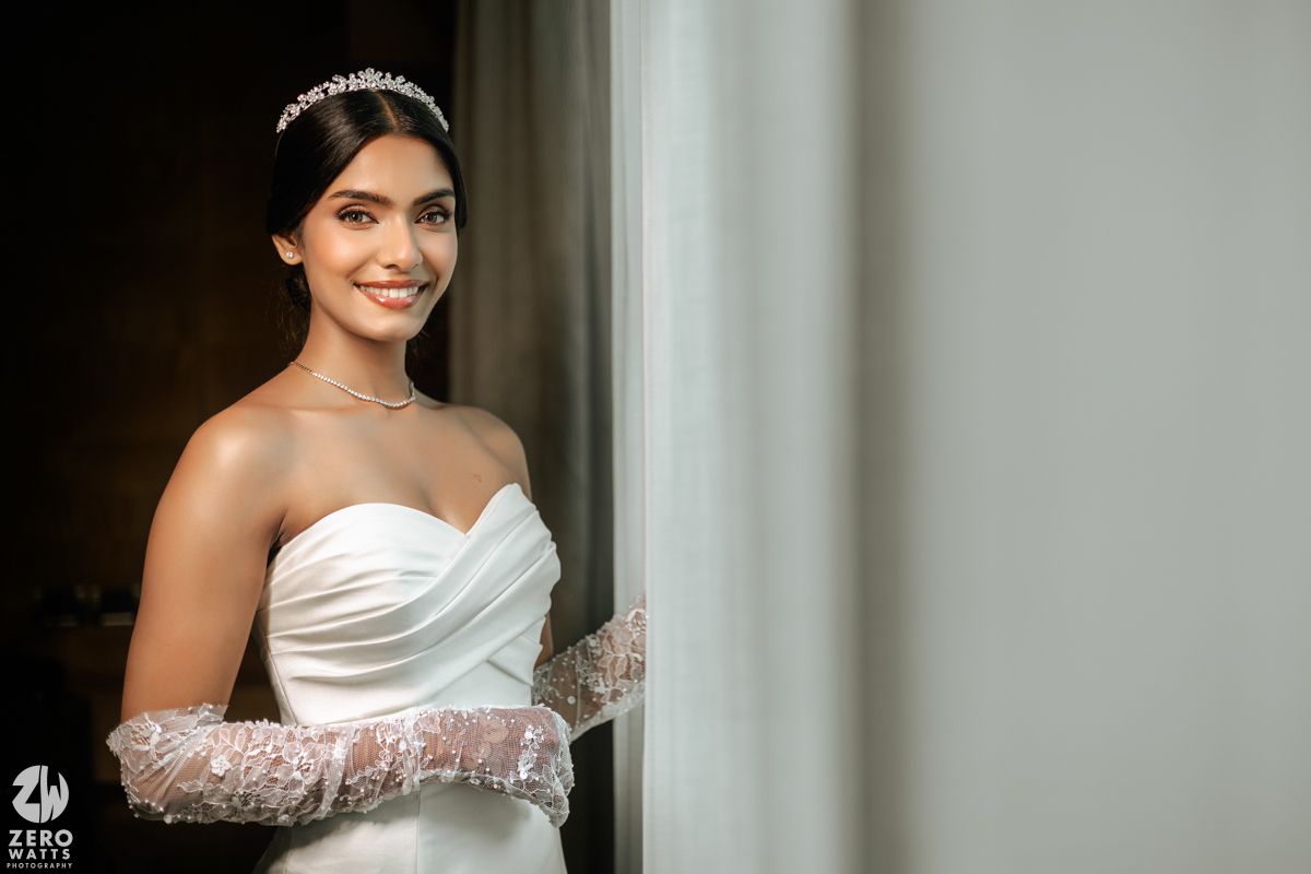 Beautiful bride wearing strapless white gown, crystal tiara, and lace gloves smiles near a window.