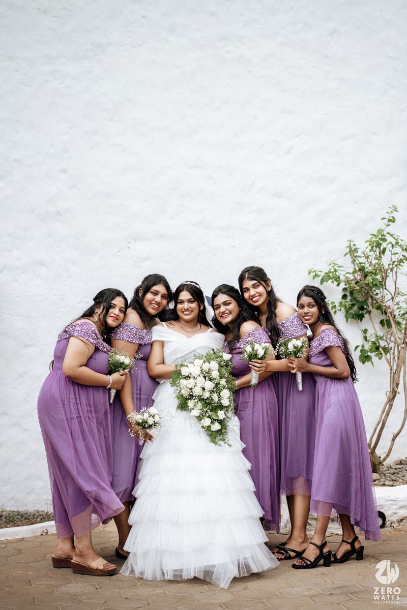 Bride in white tiered gown poses with six bridesmaids in purple dresses holding white floral bouquets.