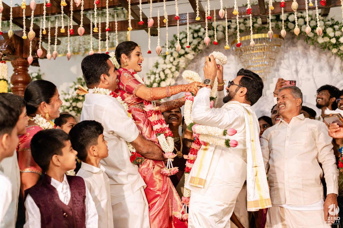 Joyful South Indian wedding garland exchange ceremony with groom lifted by groomsmen amid floral decorations.