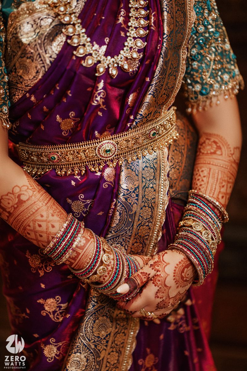 Close-up of Indian bride wearing purple saree with gold jewelry, mehndi-covered hands, and colorful bangles.