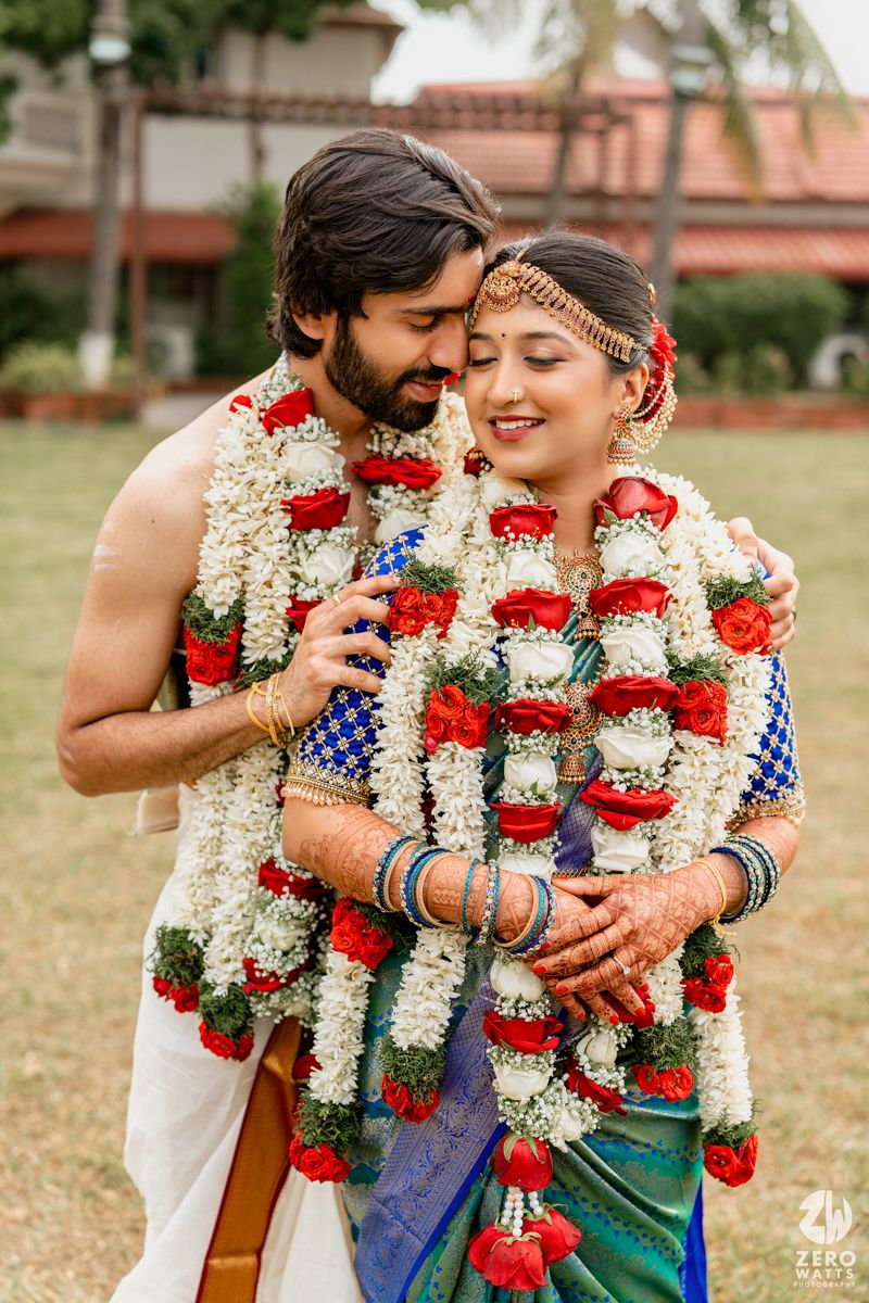 Indian bride and groom in traditional attire embracing, adorned with red and white wedding garlands outdoors.