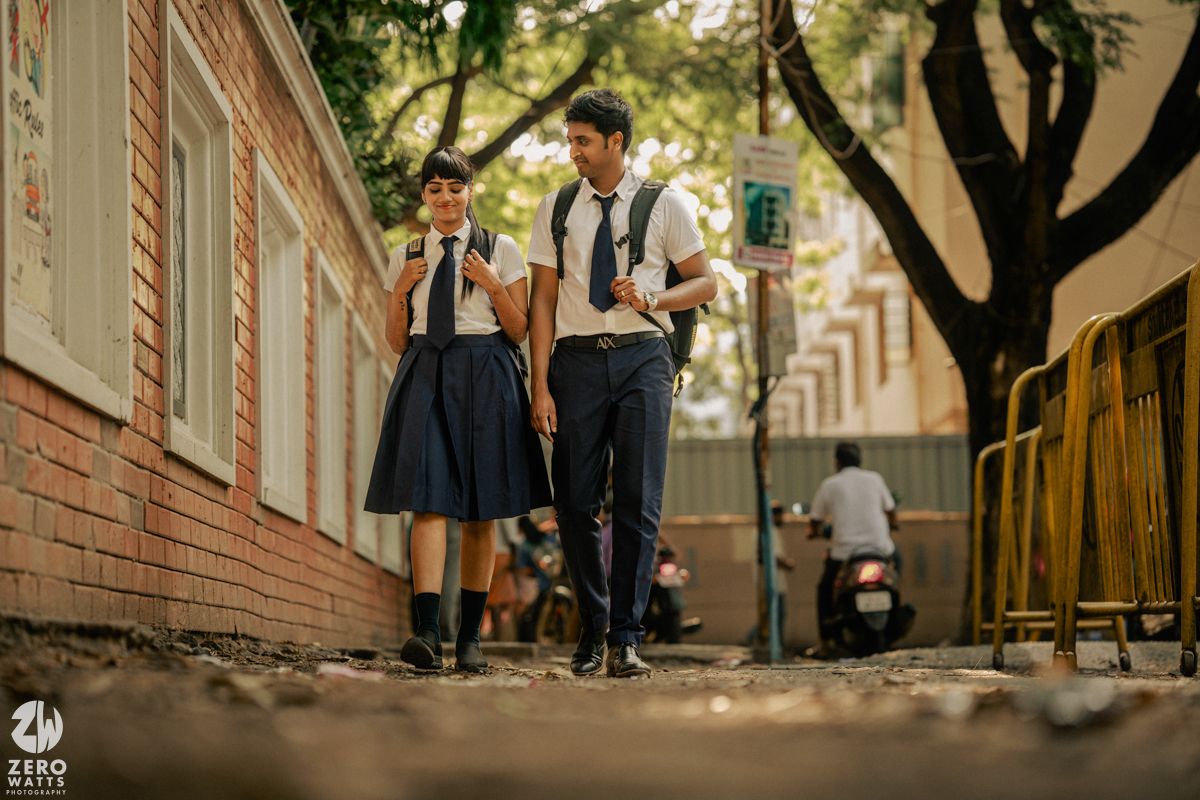 A vintage-styled couple in school uniforms stands together on a tree-lined street with fallen leaves.