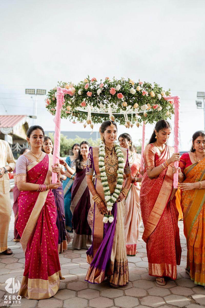 Group of women in colorful sarees celebrating at outdoor wedding venue under floral arch decorations.