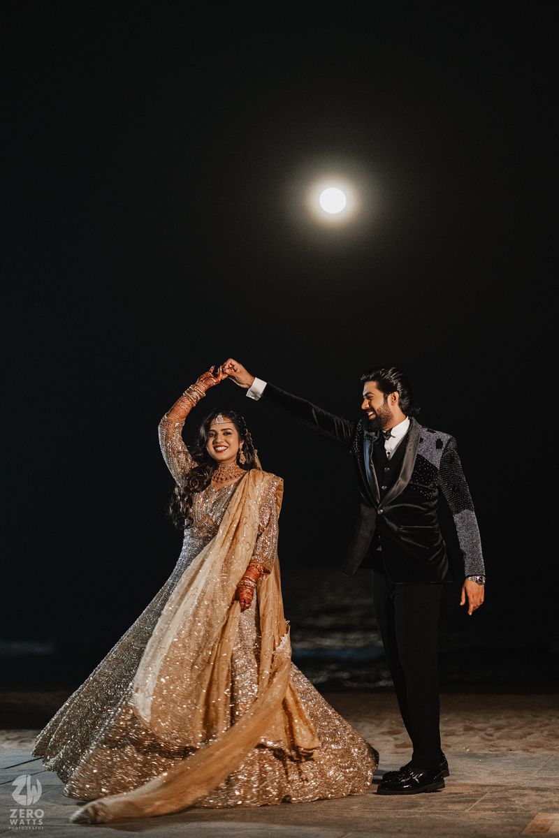 Elegant wedding couple poses on a dark beach with flowing sequined lehenga and veil creating magical silhouettes.