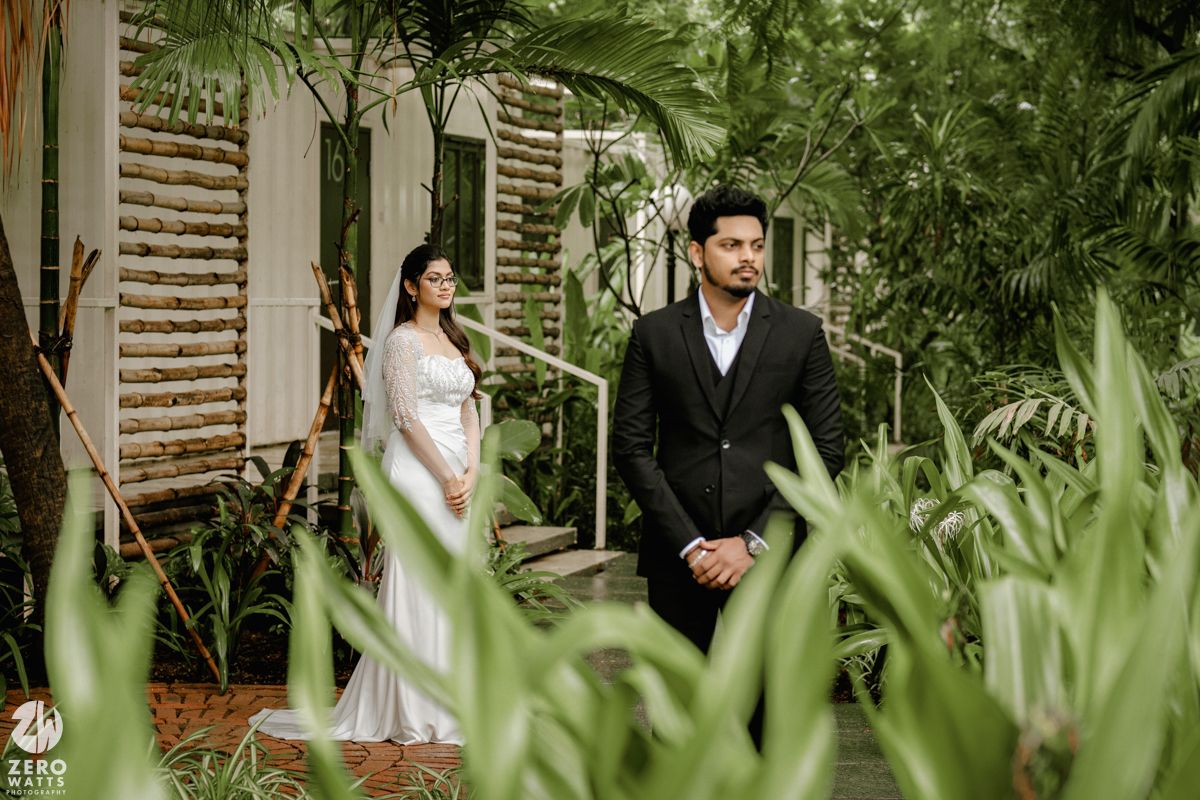 Wedding party stands in lush tropical garden setting with wooden wall and green foliage in background.