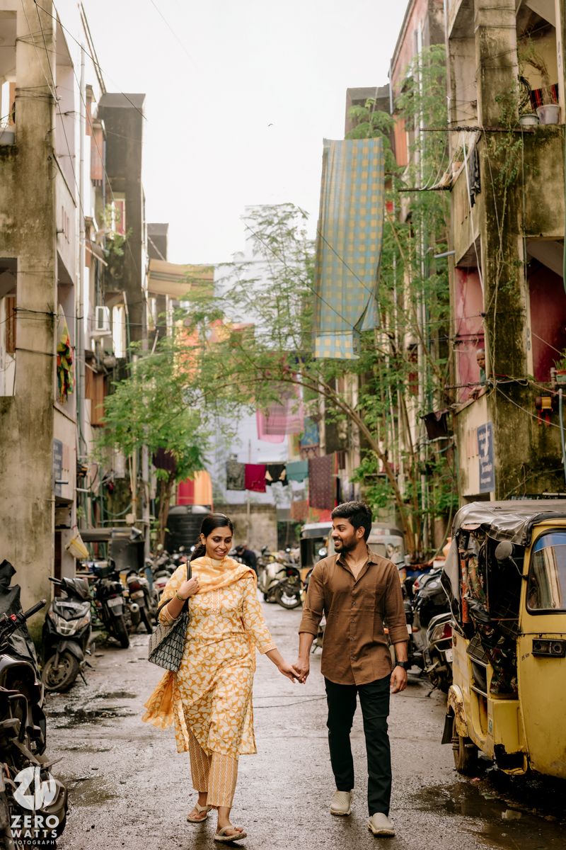 A couple walks hand in hand down a narrow urban street lined with parked motorcycles and auto rickshaws in India.