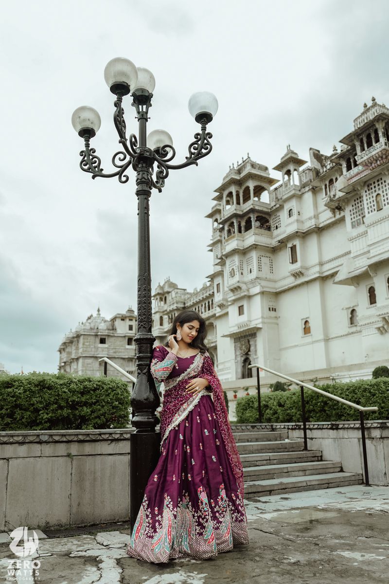 A purple and silver embroidered saree photographed against the white facade of Udaipur City Palace with an ornate lamppost.