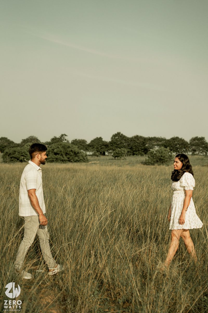 Couple walking through tall grass field with trees in background during sunset.