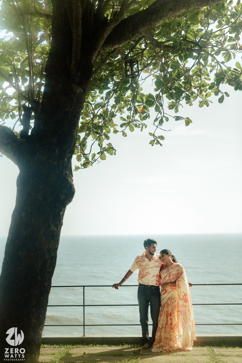 Couple in matching outfits sharing romantic moments under trees by the ocean during a sunset photoshoot.