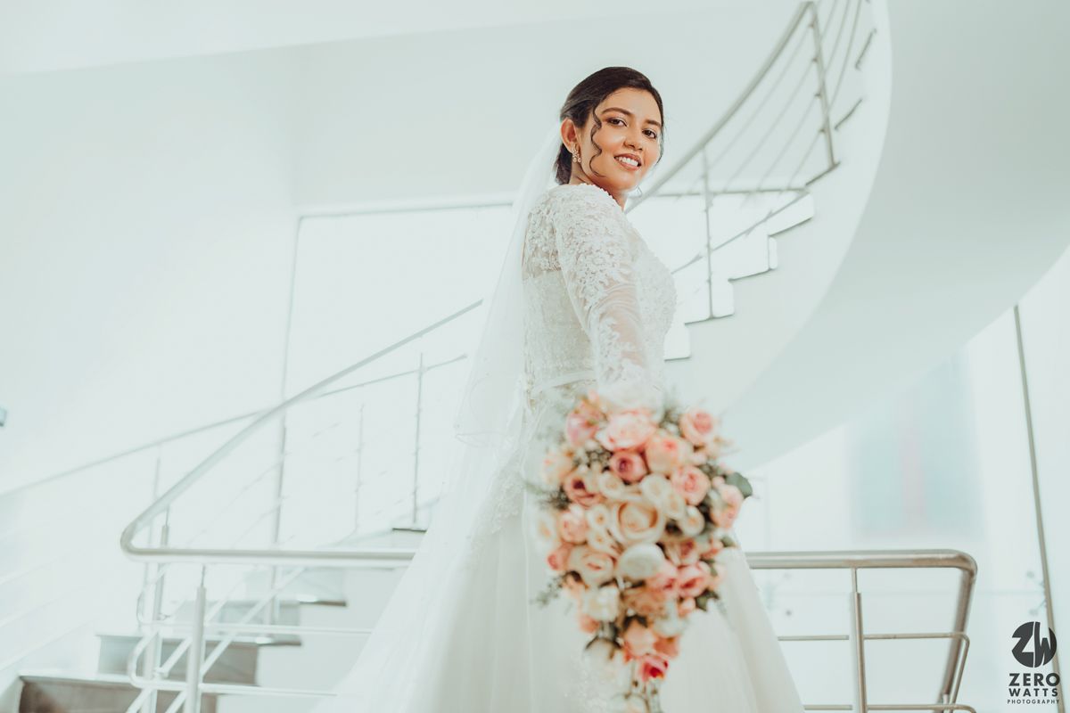 Elegant bride in a white lace gown poses on a modern white staircase with pink floral bouquet.