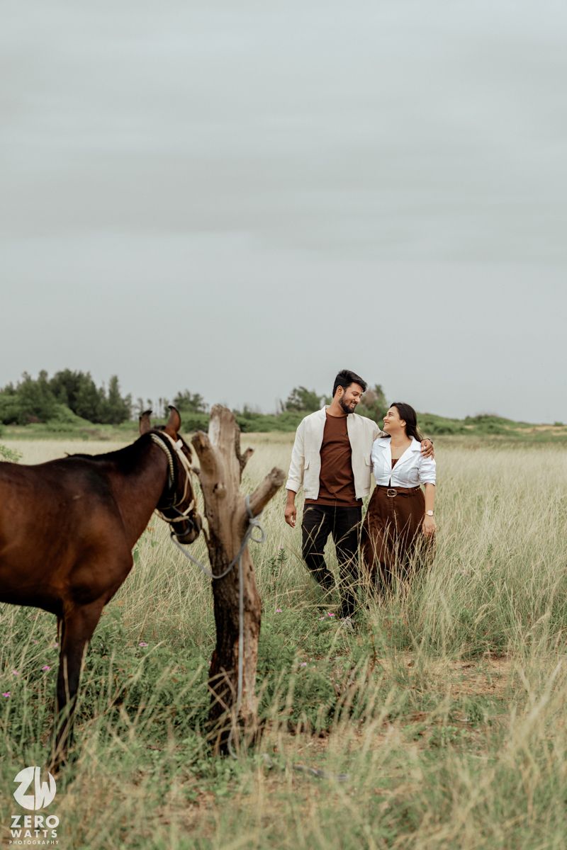 Couple embraces in a rustic field with brown horses grazing in tall grass under an overcast sky.