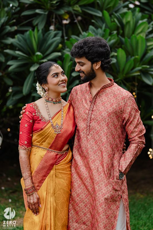 A couple in traditional South Indian wedding attire pose together against a green garden backdrop.