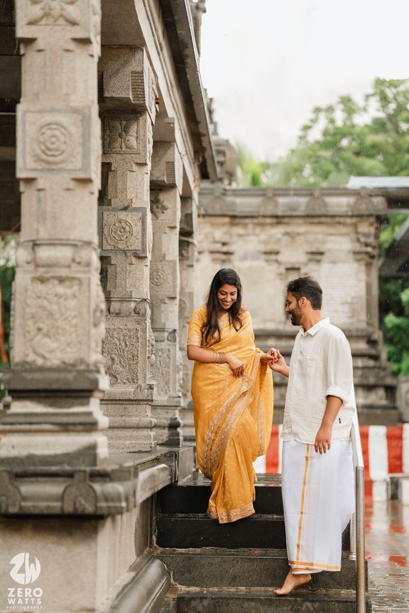 A couple in traditional Indian attire stands on stone temple steps during a rainy day photoshoot.