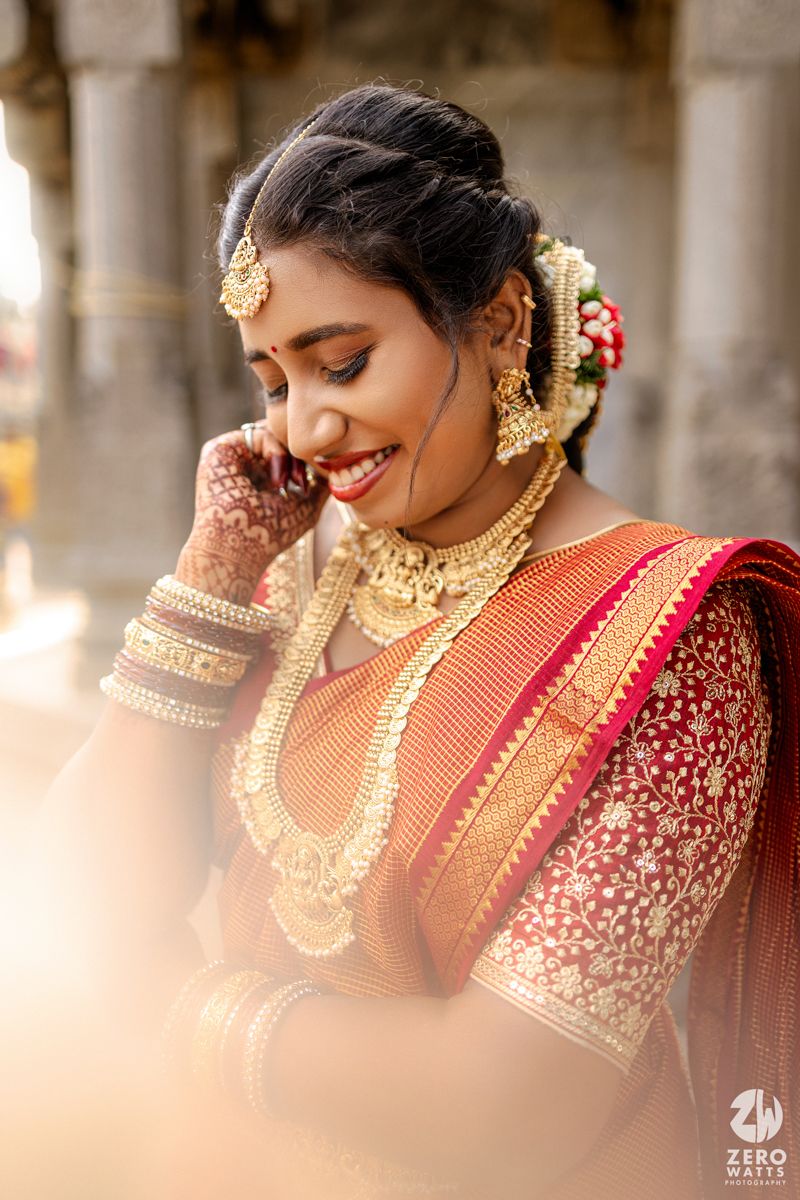 Traditional Indian bride wearing red silk saree and gold jewelry smiles while looking down in elegant wedding portraits.