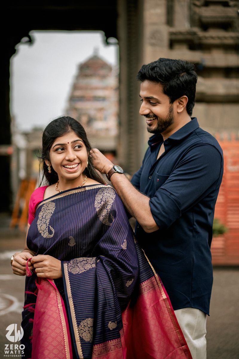 A couple shares playful moments while adjusting traditional Indian clothing against a rustic backdrop.