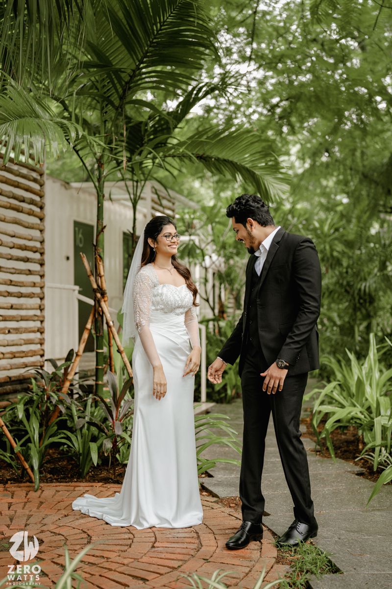 A bride and groom share a romantic moment in a lush tropical garden setting with brick pathways and palm trees.