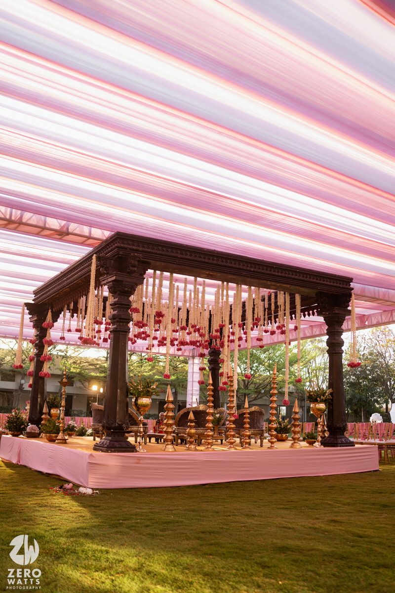 Elegant dark wood mandap adorned with hanging gold bells and floral decorations under a pink striped wedding tent.
