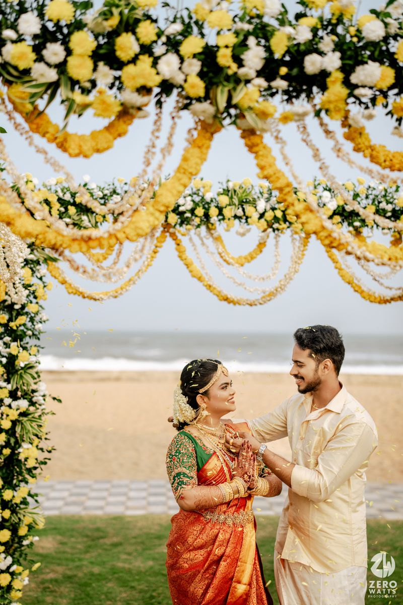 Bride in orange saree and groom in cream kurta under yellow floral beach wedding mandap.