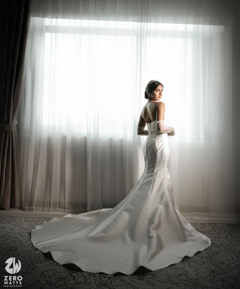 Bride in elegant backless white satin mermaid wedding gown with long train standing by sheer curtained window.