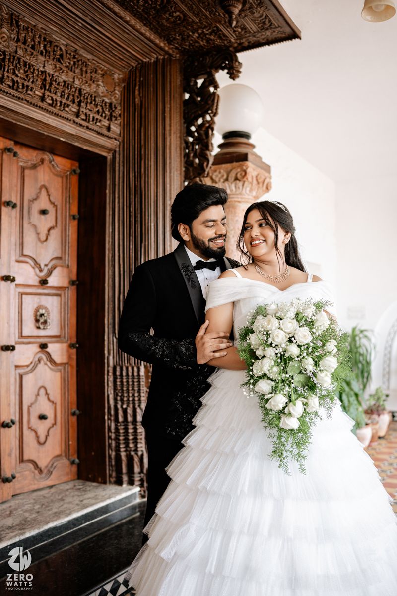 Bride in white ruffled gown holds bouquet while groom in black tuxedo embraces her near ornate wooden door.