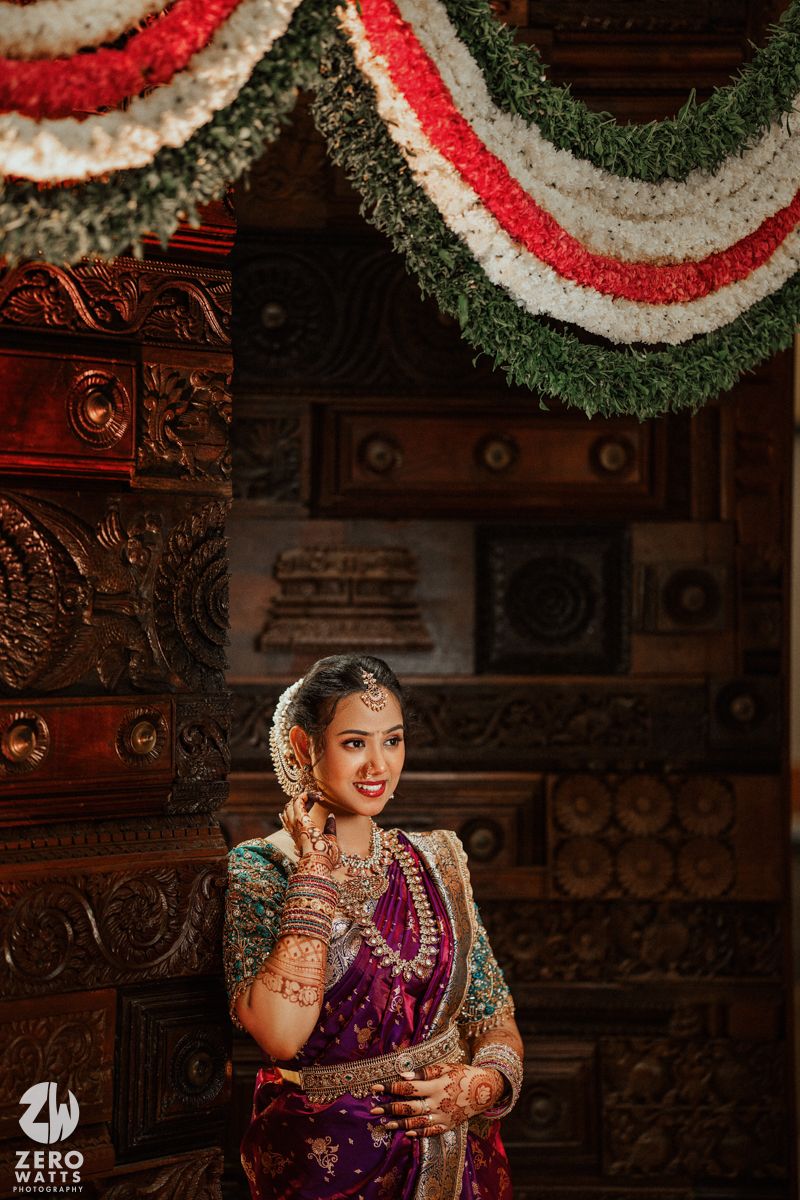 South Indian bride in purple silk saree with gold jewelry poses under floral garlands at ornate wooden temple backdrop.