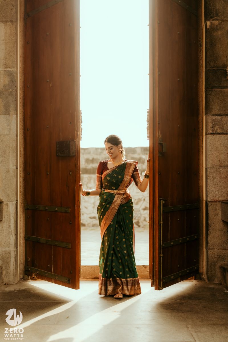 Woman in elegant green and gold silk saree standing in sunlit doorway of ancient stone building.