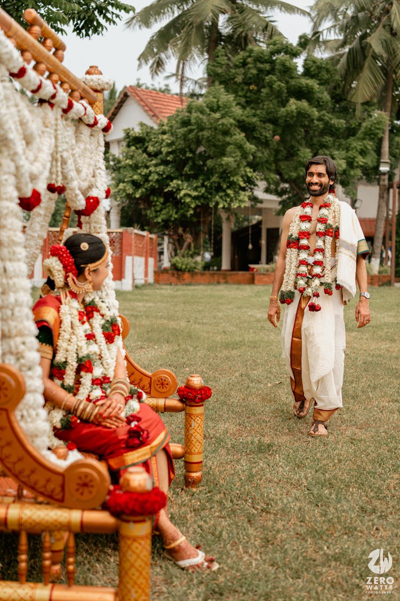 A groom in white dhoti walks toward a bride in red saree seated at a floral mandap during a South Indian wedding ceremony.