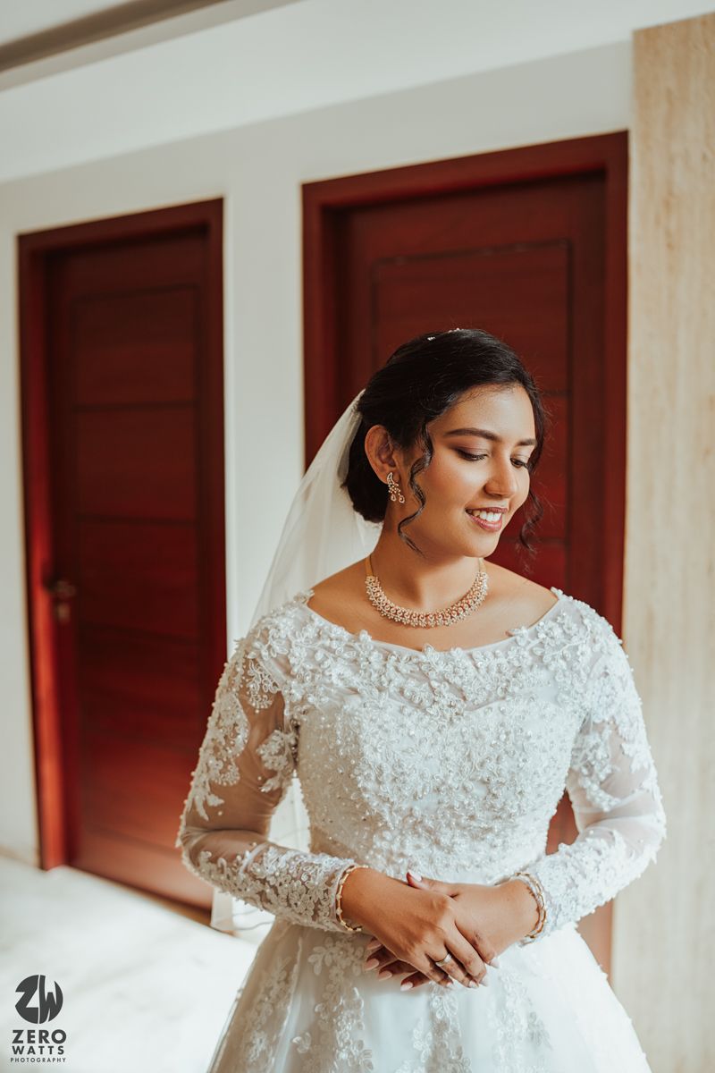 A bride wearing an elegant white lace wedding gown with long sleeves poses joyfully against a red door backdrop.