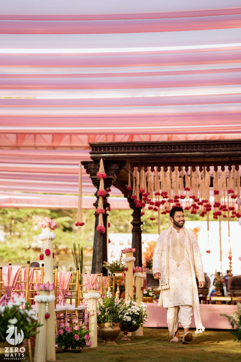 Groom in white sherwani stands at decorated Indian wedding mandap with pink draping and floral arrangements.
