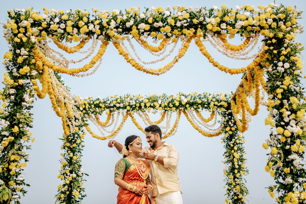 Indian couple in traditional attire posing under a yellow floral arch decorated with marigolds and white flowers at a wedding.