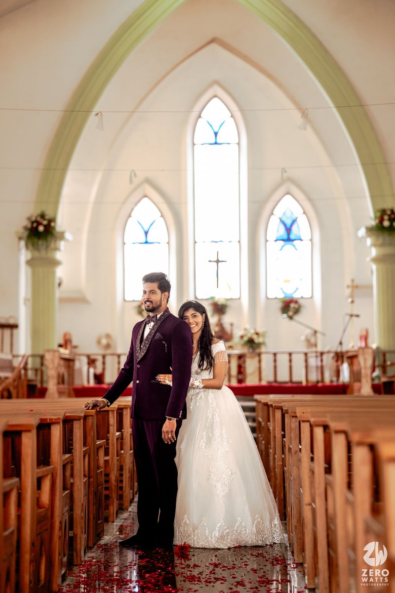 Bride and groom posing elegantly in a white church aisle, groom in purple suit, bride in lace wedding gown.