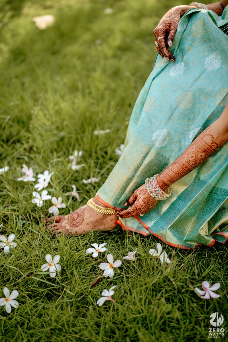Bride in teal saree with intricate mehndi on hands and feet, gold anklet, seated on grass with white flowers.