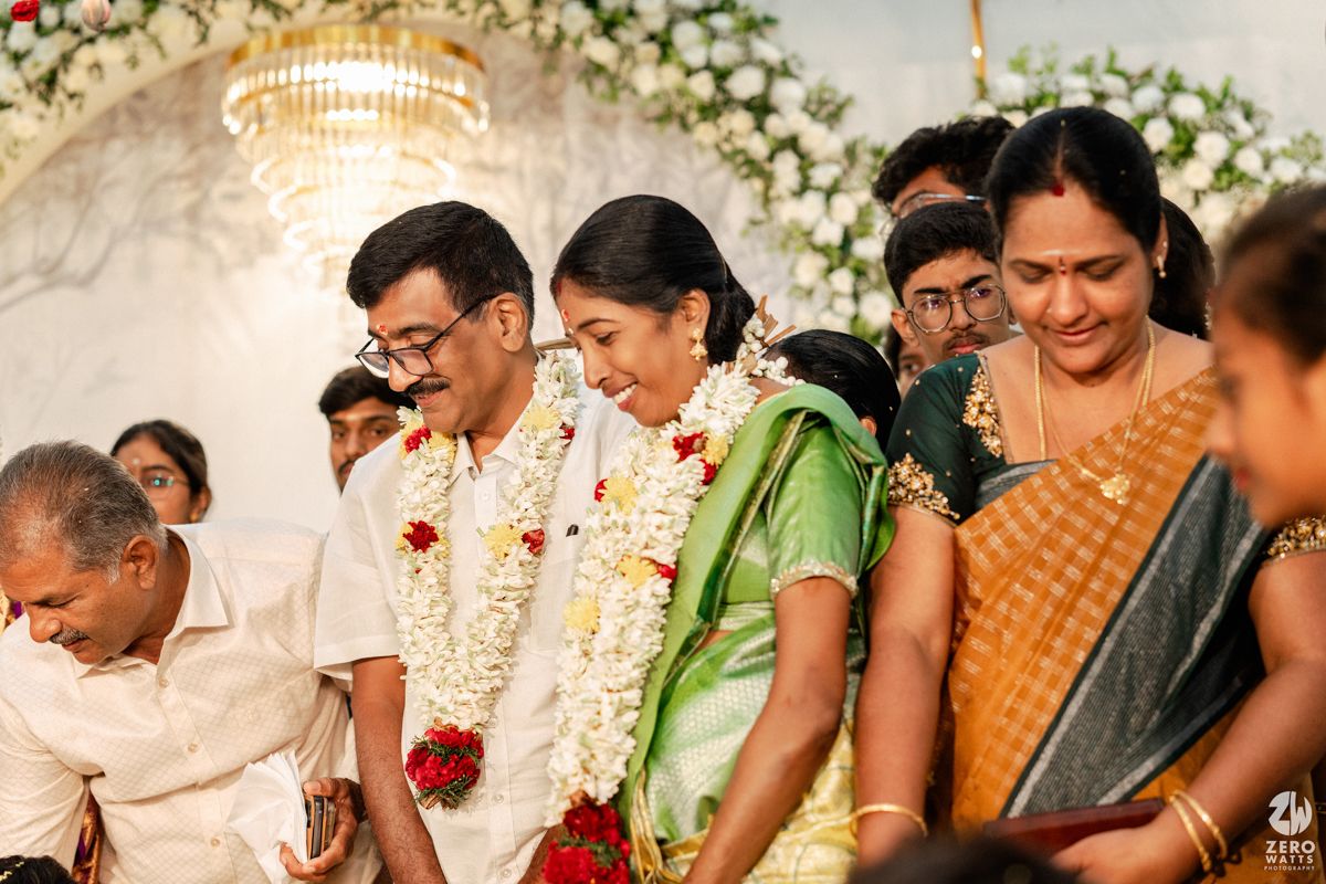 South Indian wedding ceremony with couple exchanging garlands under chandelier with family present.