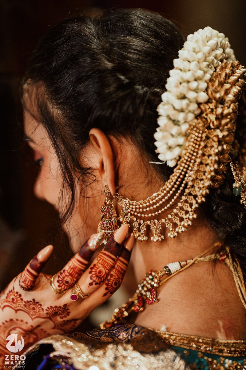 Close-up of Indian bride with mehndi hands adjusting gold necklace, white flowers in dark hair.