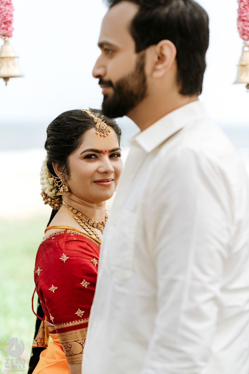 South Indian bride in red and orange saree with gold jewelry gazes at groom during outdoor wedding.