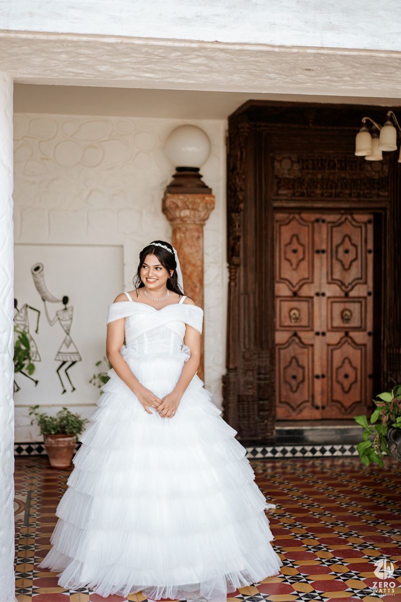 Bride in white off-shoulder tiered ball gown standing before ornate wooden doors on colorful tiled floor.