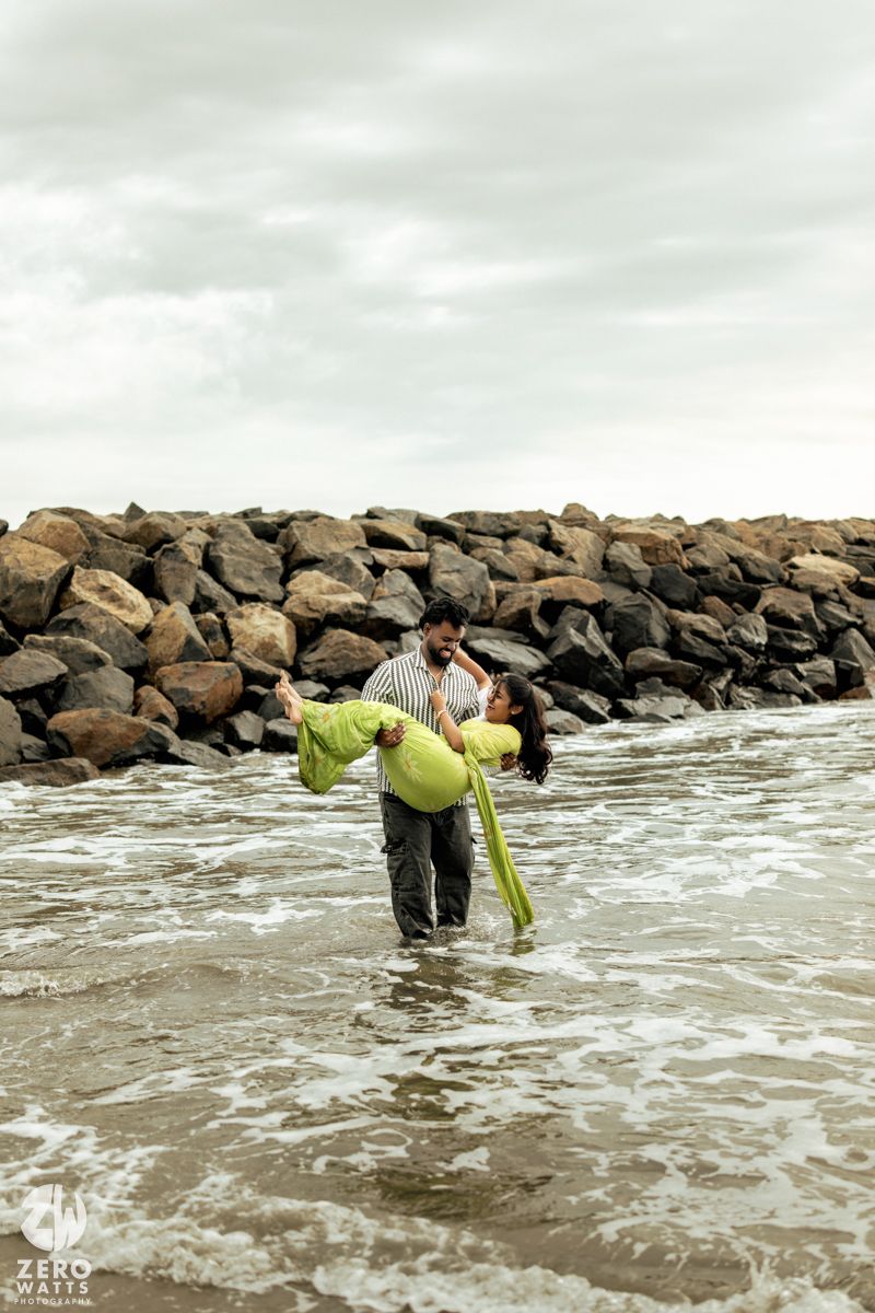 Two people wade through ocean waves near a rocky breakwater during a coastal photoshoot.