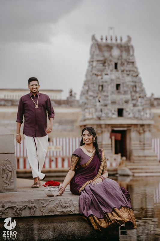 A couple in purple and white outfits pose playfully near a traditional temple water feature.