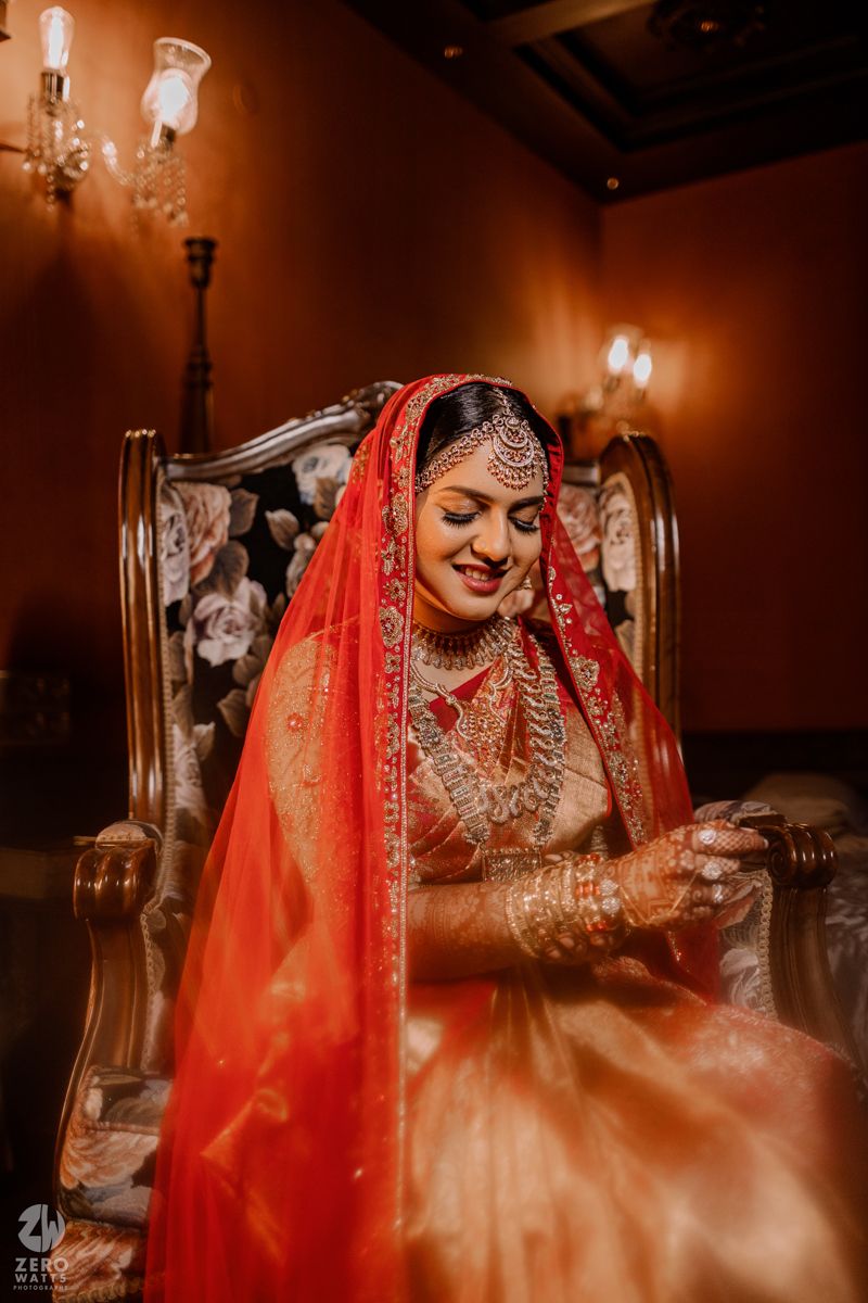 A bride in a red traditional Indian wedding dress and veil sits in an elegantly lit room.